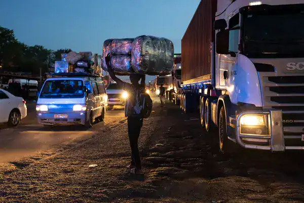 Person carrying a large bag walking past trucks and cars on a busy road at dusk in Cape Town.