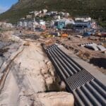 Wide aerial view of stepped concrete revetment construction along Muizenberg beachfront with machinery, beach and buildings below the mountain.