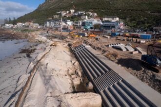 Wide aerial view of stepped concrete revetment construction along Muizenberg beachfront with machinery, beach and buildings below the mountain.