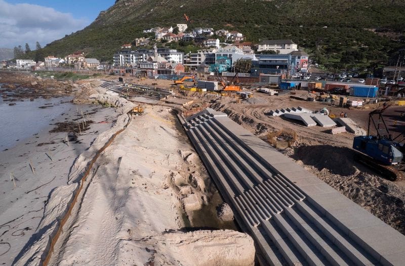 Wide aerial view of stepped concrete revetment construction along Muizenberg beachfront with machinery, beach and buildings below the mountain.